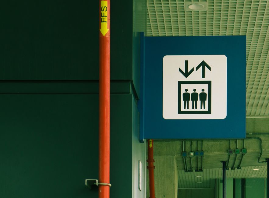 A blue and white sign displaying an elevator with an icon of three people inside, indicating the location of passenger lifts in a building. The sign features upward and downward arrows, signifying the direction of travel within the elevator. It is mounted on a wall near a corridor ceiling, adjacent to the green wall section on the left and beneath a ceiling with a grid pattern. Several visible pipes and conduit are mounted on the wall below the sign. This signage is part of the building's internal wayfinding system, essential for guiding residents or visitors during home relocation or moving processes. The environment appears to be an indoor area likely within a multi-story building, supporting logistics and transport arrangements for furniture and belongings during house removals facilitated by companies such as Man with Van West Norwood.
