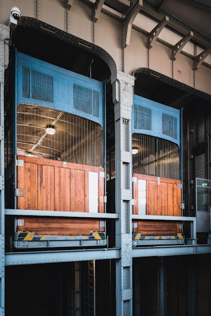 Interior view of a building with two large freight elevator shafts designated for home relocation and furniture transport in West Norwood, featuring wooden loading platforms secured with metal brackets and transparent safety panels. The elevators are built with sturdy grey metal frames and black rubber bumpers along the edges, with the upper sections revealing a high ceiling and industrial lighting. The environment is illuminated with artificial light, highlighting the structural components and the secure loading areas used for moving furniture, boxes, and appliances. This setting exemplifies the logistical considerations involved in packing and moving processes, such as the use of lifts for efficient bulk transport during residential relocations, with the professional services of Man with Van West Norwood potentially assisting with planning and executing such home moves.