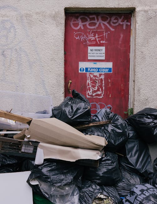 A pile of black garbage bags and discarded packaging materials, including cardboard boxes and plastic sheeting, stacked against a textured, off-white exterior wall near a weathered red metal door covered with graffiti and various signs, including one reading 'Keep clear' and another indicating 'No money.' The bags and materials are placed on the pavement, close to the doorway, suggesting a waste disposal or clearance process. The door is partially open at the top, revealing a dark interior space, and the area shows signs of neglect typical for an urban setting. This scene reflects a typical debris removal task that could be associated with house or furniture clearance as part of moving or relocation services offered by Man with Van West Norwood, in the context of home relocation or furniture transport preparations.