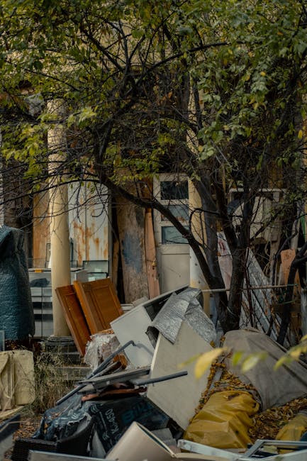An outdoor scene showing a pile of household rubbish and discarded items beneath a tree, with branches and green leaves overhead. The waste includes wooden door panels, a white ceramic sink wrapped in protective plastic, a grey fabric-covered item, and various cardboard boxes and plastic bags containing rubbish. The debris is scattered on the ground in front of a weathered fence and a building with a partially visible window and peeling paint. A large tree with dark, twisting branches provides partial shelter over the clutter. In the background, there are glimpses of a residential area, and the overall scene appears to be an untidy outdoor space awaiting clearance. This image highlights waste collection typically associated with bulky rubbish removal during house clearance or relocation, as managed by Man with Van West Norwood while preparing for efficient home relocation logistics.