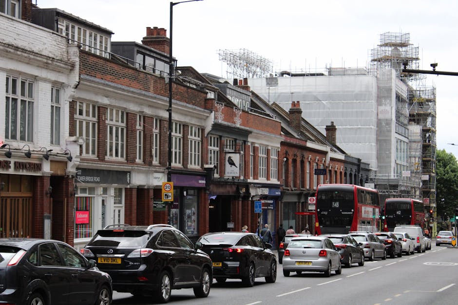 A row of two-story brick and plaster residential buildings along a busy urban street with retail shops on the ground floor, some with signage, and multiple parked and moving vehicles including black and silver cars and red double-decker buses. The street is lined with parked cars on one side, and pedestrians are walking on the sidewalk in front of the shops. Behind the buildings, there is a construction site with scaffolding and white protective sheeting covering part of a building under renovation. The scene is captured during daylight with natural lighting, illustrating a typical east London street scene relevant to house removals and furniture transport services, as managed by Man with Van West Norwood, incorporating elements of loading and moving logistics within an active city environment.