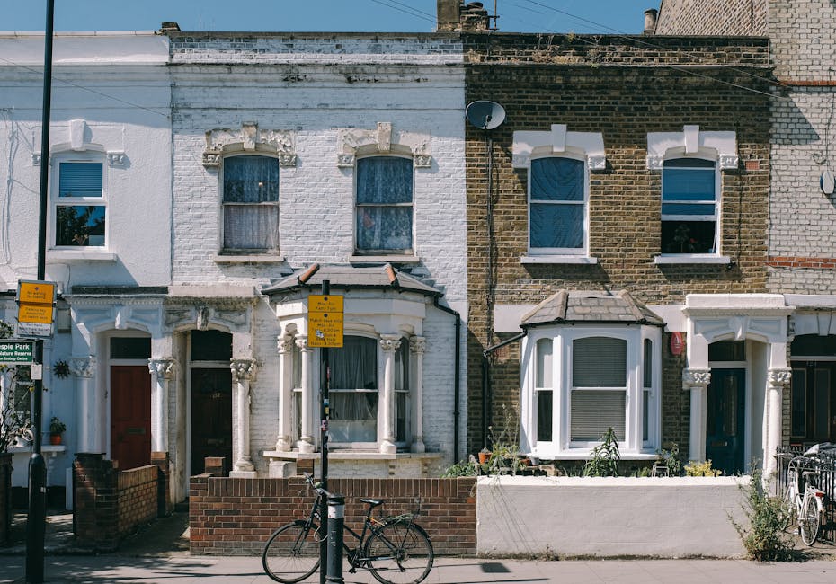 A row of terraced houses with traditional bay windows and brick facades, situated on a street with a bicycle parked against a brick wall in front. The house on the left has a white painted exterior with decorative stone details around the windows and a small front garden enclosed by a low brick wall. The house on the right has a darker brick exterior with a similar bay window and garden area, also enclosed by a low wall. In front of the houses, there are two traffic signs mounted on black poles and a bicycle resting against a brick boundary wall, with a clear blue sky overhead. The scene captures typical residential street environment, suitable for house removals and home relocation services by companies like Man with Van West Norwood, with visible features indicating living spaces and street-level logistics involved in furniture transport and packing during moving processes.