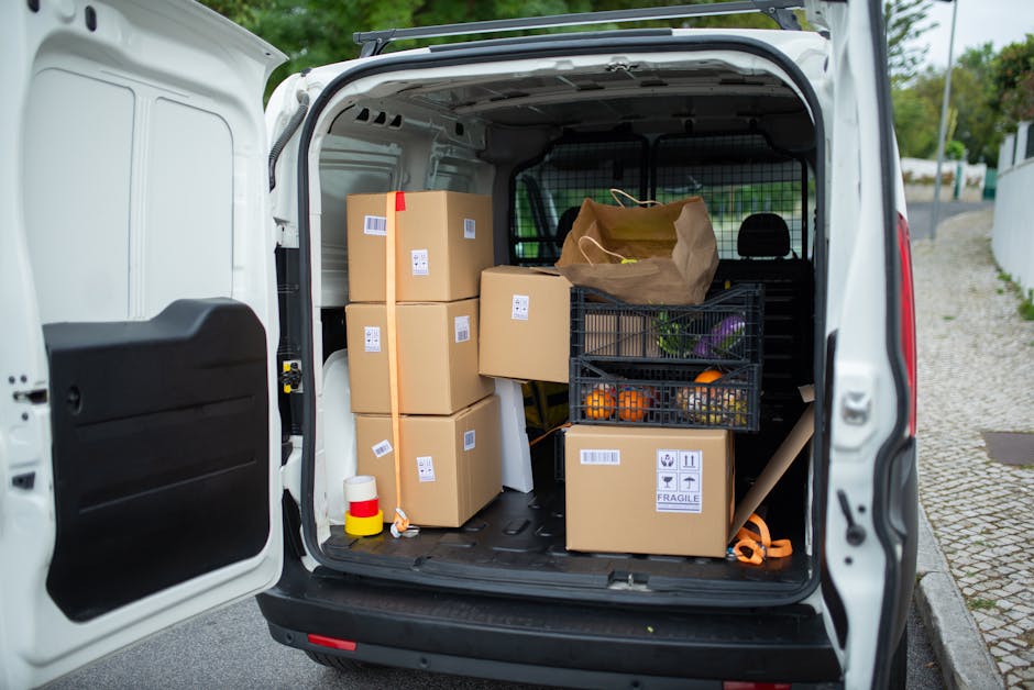 Inside the open rear cargo area of a white van parked on a street, several cardboard boxes of various sizes, some sealed with tape and labeled for fragile contents, are stacked neatly. A black plastic crate contains fresh vegetables, and a brown paper shopping bag is placed atop one of the boxes. The van's interior shows a black protective partition behind the cargo space, and a yellow and red safety cone is positioned on the floor beside the boxes. The surroundings include cobbled pavement and a white fence, with greenery and a partly cloudy sky visible in the background. This scene depicts a home relocation or furniture transport process, with items prepared for moving services offered by Man with Van West Norwood, aligned with busy packing and loading activities during a house move.