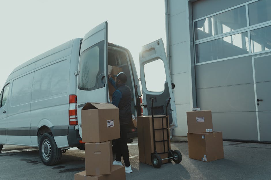 A man wearing a dark jacket and a cap is loading cardboard boxes into the rear of a white moving van parked outside a modern building with large glass windows and a metal facade. The boxes are made of brown corrugated cardboard, some with packing labels, and are being carried on a hand truck with black handles and small wheels. The van's side door is open, revealing additional packed boxes and moving supplies inside. The loading action takes place on an outdoor paved surface, illuminated by natural daylight, with the sun visible behind the building. Several sealed cardboard boxes are placed on the pavement nearby, ready for transport as part of a home relocation or furniture transport process. The scene depicts professional removals work, likely carried out by a company such as Man with Van West Norwood, to facilitate packing and moving services in the SE27 area.