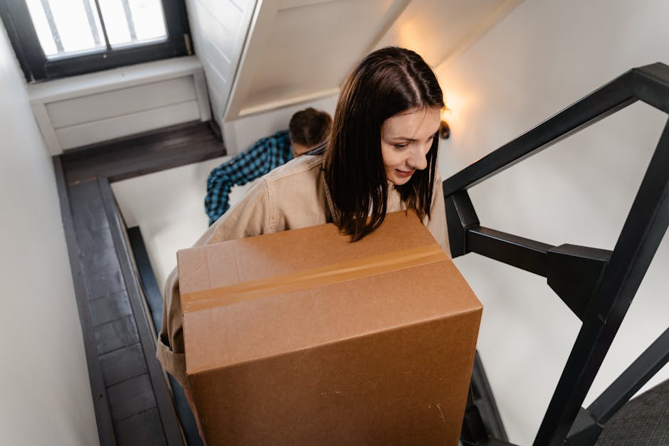 A woman with dark brown hair, wearing a beige jacket, is carrying a large cardboard box up a narrow staircase inside a residential property. Behind her, a young man with short hair in a blue and black checked shirt is also ascending the stairs with a box, partially visible. The staircase has dark grey treads and white risers, with a black metal railing on the right side, and the stairs lead to an upper floor with a white wall and a window at the top. Natural light from the window illuminates the scene, highlighting the plastic wrap and packing materials on the boxes, which are part of a home relocation process. The image captures the loading process, including lifting and carrying of boxes, typical of furniture transport and packing during house removals, as executed by Man with Van West Norwood, specialists in removals and moving services.