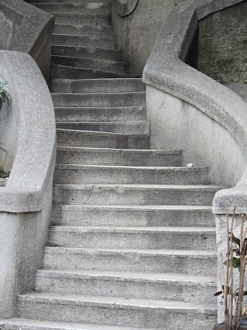 A wide, curved stone staircase with multiple steps leading upward, featuring smooth, rounded handrails on each side that extend from the bottom to the top. The stairs are made of grey concrete or limestone and show signs of weathering. On the right side near the lower steps, a small bundle of dried branches is visible. The staircase is situated outdoors, framed by stone walls on either side, and appears to be part of a historic or decorative garden feature. The lighting suggests natural daylight, with no visible furniture, boxes, or moving equipment present. This setting reflects a typical exterior staircase which can be a challenging feature for home relocations involving bulky furniture or large items that need to be navigated through narrow or curved stairways, an area where [COMPANY_NAME] offers professional removal solutions and furniture transport services.