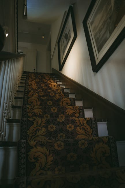 A woman with dark brown hair, wearing a beige jacket, is carrying a large cardboard box up a narrow staircase inside a residential property. Behind her, a young man with short hair in a blue and black checked shirt is also ascending the stairs with a box, partially visible. The staircase has dark grey treads and white risers, with a black metal railing on the right side, and the stairs lead to an upper floor with a white wall and a window at the top. Natural light from the window illuminates the scene, highlighting the plastic wrap and packing materials on the boxes, which are part of a home relocation process. The image captures the loading process, including lifting and carrying of boxes, typical of furniture transport and packing during house removals, as executed by Man with Van West Norwood, specialists in removals and moving services.