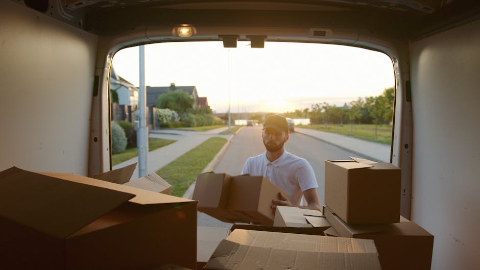 A person inside a moving van, seen from the rear open door, is loading cardboard boxes onto the vehicle during a home relocation. The individual, wearing a cap and white shirt, is handling several medium-sized boxes, some with flaps open, indicating packing and moving preparations. The van's interior is spacious, with a few packing materials visible, and the boxes are situated on the floor near the tailgate, ready for transport. Outside the van, a suburban street in West Norwood is visible, illuminated by sunlight, with a sidewalk, lawn, trees, and residential houses in the background. The scene captures the logistical process of furniture transport and packing during house removals, typical of [COMPANY_NAME]'s moving services in the SE27 area, as part of a scheduled home move on Norwood Road.