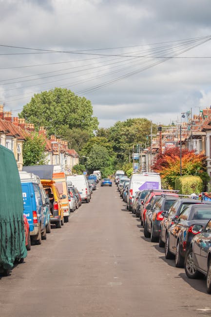 A residential street scene shows a typical suburban neighbourhood with terraced houses on both sides, each with small front gardens and a variety of trees and shrubs in full leaf. Parked along both sides of the narrow tarmac road are numerous cars and vans, including white, blue, and grey vehicles, some of which appear to be commercial or moving vans. In the foreground, a man with a van service, associated with Man with Van West Norwood, is seen unloading or loading furniture and moving boxes covered with plastic wrap and cardboard packaging from a large white van into the property. The furniture includes a wooden chest of drawers and a cardboard box on a trolley, with items being carefully carried into the house through the front door. The scene is set during daytime with overcast skies and soft natural light, illustrating a typical home relocation process involving furniture transport and packing and moving activities, crucial steps in a house removal in the SE27 area of West Norwood.