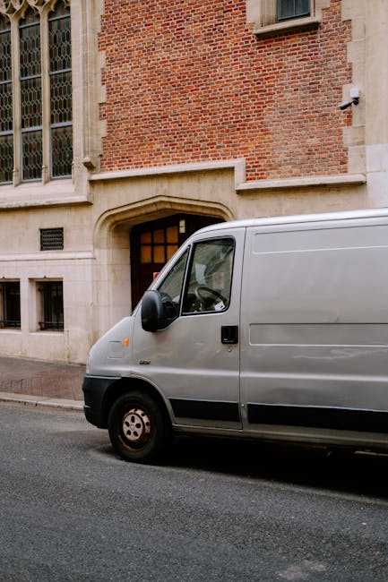 A silver moving van parked on the street in front of a historic brick building with large stone window sills and tall windows. The van is positioned close to the building's doorway, with its side facing the camera. The loading area of the van is not visible, but the vehicle’s front passenger door is closed, and there are no people in the image. The street appears to be quiet and clean, with a grey pavement surface. This scene depicts the initial stage of a home relocation or furniture transport process, as part of professional removals services by Man with Van West Norwood, supporting the logistics of packing, loading, and moving household items. The building’s architectural features include stone details around the windows and an entrance with an arched stone surround, indicating a residential or institutional property typical in the Norwood Road area. The image emphasizes the preparation and planning involved in house removals, with the van positioned for efficient loading or unloading, all under natural daylight conditions.