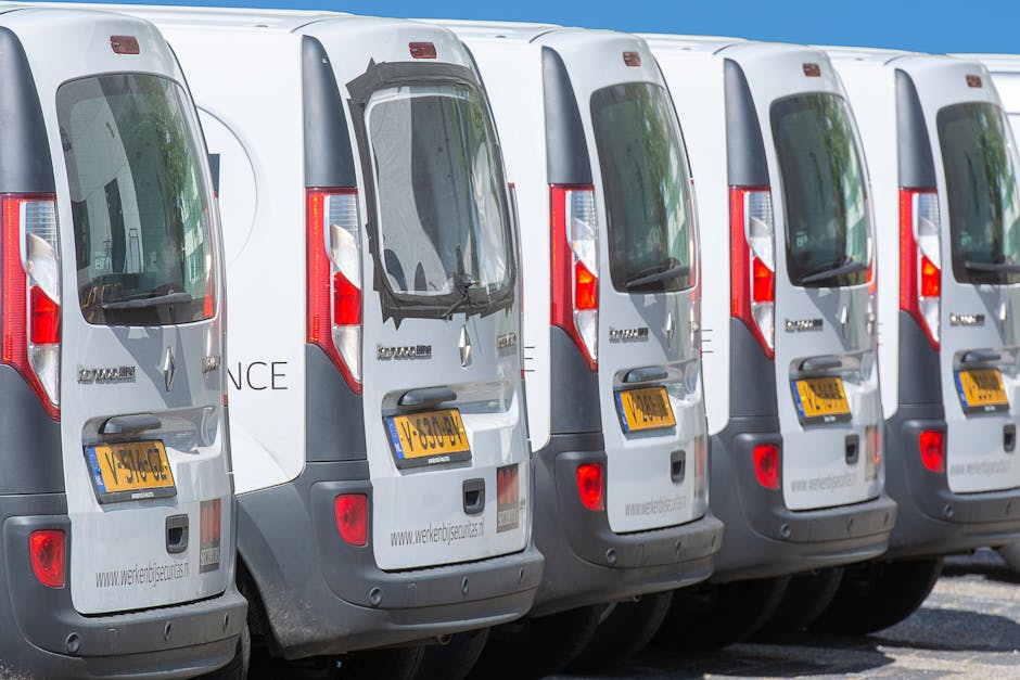 A row of five white commercial vans parked closely together on a street, with their rear doors and license plates visible. The vans are positioned side by side, facing away from the camera, with a clear view of their back windows, taillights, and rear bumpers. The vans are part of a moving or furniture transport fleet used for house removals, with the side of one van displaying the company's website, indicating professional relocation services. The environment is outdoors with bright daylight, and the vans are situated on a paved surface. This setup reflects the logistical aspect of home relocation or furniture transport, handled by [COMPANY_NAME], supporting efficient loading and unloading processes for house removals.