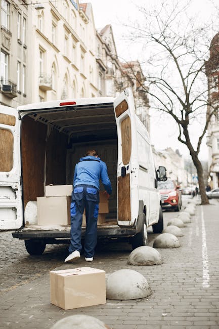 A silver moving van parked on the street in front of a historic brick building with large stone window sills and tall windows. The van is positioned close to the building's doorway, with its side facing the camera. The loading area of the van is not visible, but the vehicle’s front passenger door is closed, and there are no people in the image. The street appears to be quiet and clean, with a grey pavement surface. This scene depicts the initial stage of a home relocation or furniture transport process, as part of professional removals services by Man with Van West Norwood, supporting the logistics of packing, loading, and moving household items. The building’s architectural features include stone details around the windows and an entrance with an arched stone surround, indicating a residential or institutional property typical in the Norwood Road area. The image emphasizes the preparation and planning involved in house removals, with the van positioned for efficient loading or unloading, all under natural daylight conditions.
