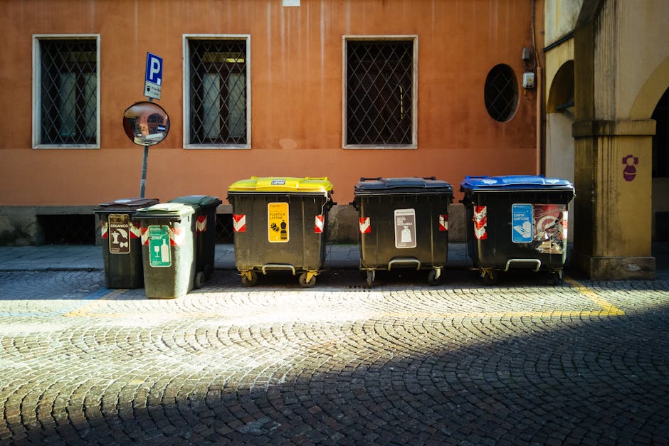 A row of four large waste and recycling bins positioned on a cobblestone pavement outside a building in West Norwood, with a peach-colored wall and three windows behind them. The bins are made of plastic, each with a different colour lid—green, yellow, black, and blue—indicating different waste categories. The green bin appears designated for general waste, the yellow for recyclable packaging, the black for non-recyclables, and the blue for paper or cardboard. The bins are equipped with wheels and are situated close to the building’s wall, which has decorative stone arches and an electrical box mounted to it. A small convex mirror is attached to a pole nearby, providing visibility for pedestrians and vehicles navigating the street. This scene reflects typical waste management during home relocation or furniture transport preparations, with the bins positioned for collection. Visible lighting suggests daylight, and the overall environment indicates an organized setting aligned with house removals and moving logistics, as managed by Man with Van West Norwood.
