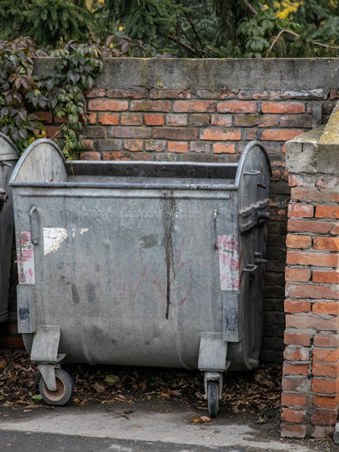 A large metal waste disposal bin with a slightly curved top and four caster wheels positioned on a pavement next to a brick wall, partially obscured by some soil and fallen leaves at the base. The bin appears weathered with visible rust, dirt streaks, and corrosion marks, and has two faded pink stickers on its side. The background features a red brick wall topped with a layer of cement, with green ivy or foliage creeping over the top. The scene is outdoors, near a residential property, with natural daylight illuminating the area. This image illustrates typical waste management in a property setting, relevant to house removals, packing, and logistics involved in residential relocations.