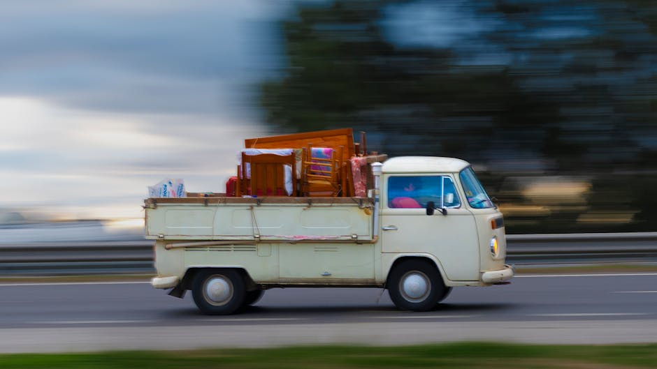 A vintage cream-colored van with a boxy shape is seen moving along a road during daylight hours, with the background appearing blurred due to motion, indicating high speed. The van's flatbed cargo area is loaded with various household furniture and items, including wooden chairs, small tables, and plastic storage containers, which are secured and stacked neatly to facilitate an efficient home relocation process. Some of these items are wrapped in protective blankets to prevent damage during transit, while cardboard boxes and plastic bags filled with packing materials are also visible, indicating a recent packing and moving activity. The scene is outdoors, with a partly cloudy sky and blurred natural surroundings suggesting the vehicle is in motion between locations. This image reflects the logistical aspect of furniture transport and packing during urgent flat moves, such as those coordinated by Man with Van West Norwood in the West Norwood area.
