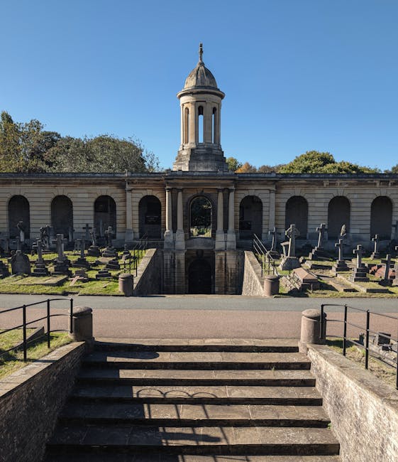 A narrow gravel pathway through West Norwood Cemetery is shaded by large trees with green and yellow leaves, casting dappled shadows on the ground. To the left of the path, several old gravestones and monuments made of stone and concrete are visible, some leaning slightly. On the right, dense foliage and trees border the path, with a glimpse of a white building or structure further along. The scene appears to be outdoors during daytime with clear, sunny weather. This setting reflects a typical site for house removals and furniture transport, where careful packing and loading may be necessary to navigate the pathway during home relocation or moving projects, with the location managed by Man with Van West Norwood.